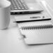 Black and white image of an open notepad next to a computer and a paperclip and pencil lie on top of the notepad